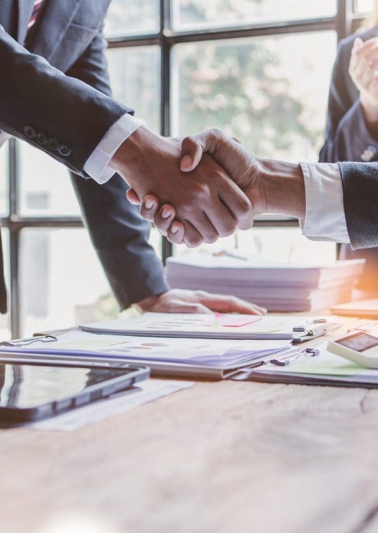 Two business professionals shaking hands over documents in an office.